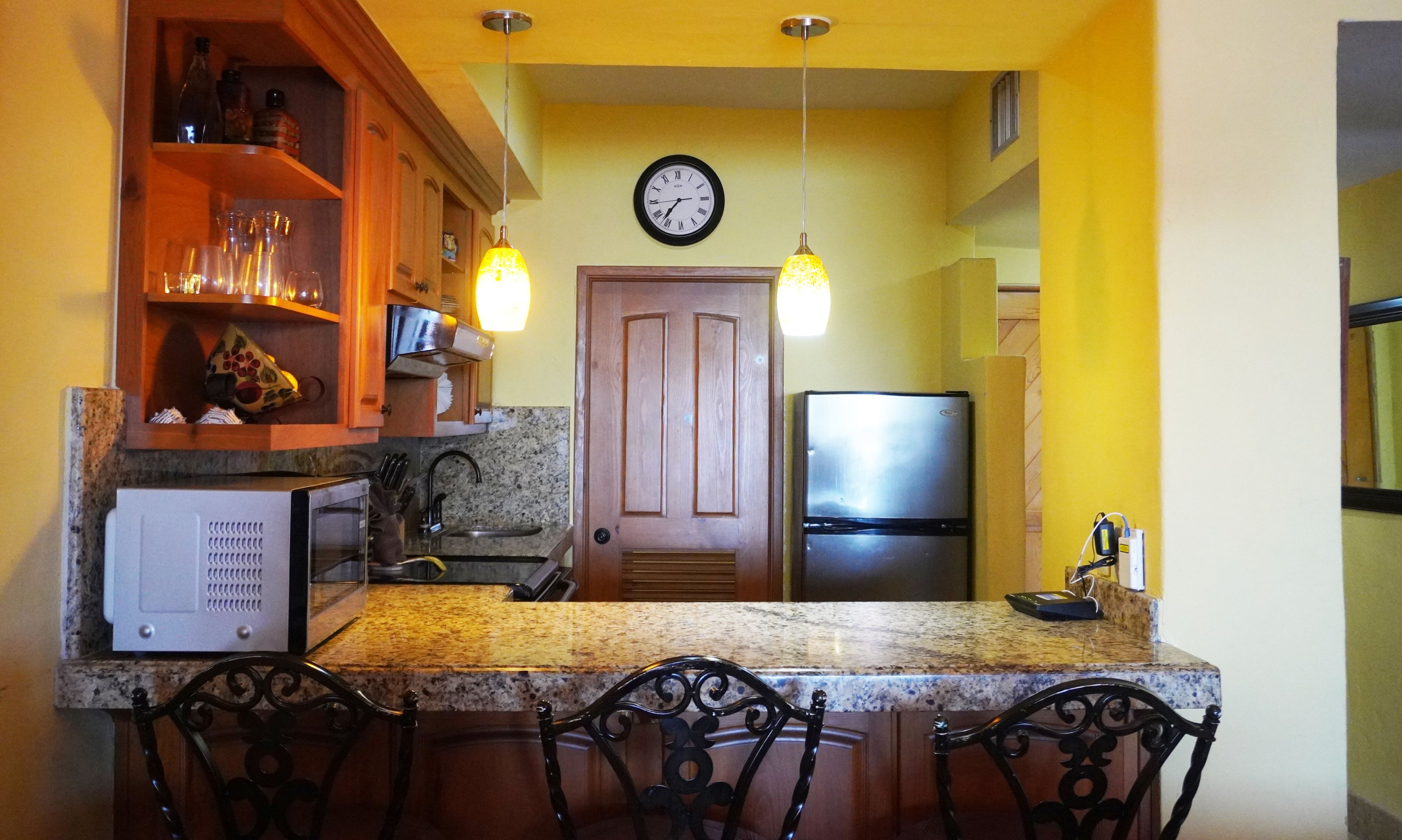 A kitchen with granite countertops, a microwave, a refrigerator, and a clock on the wall.