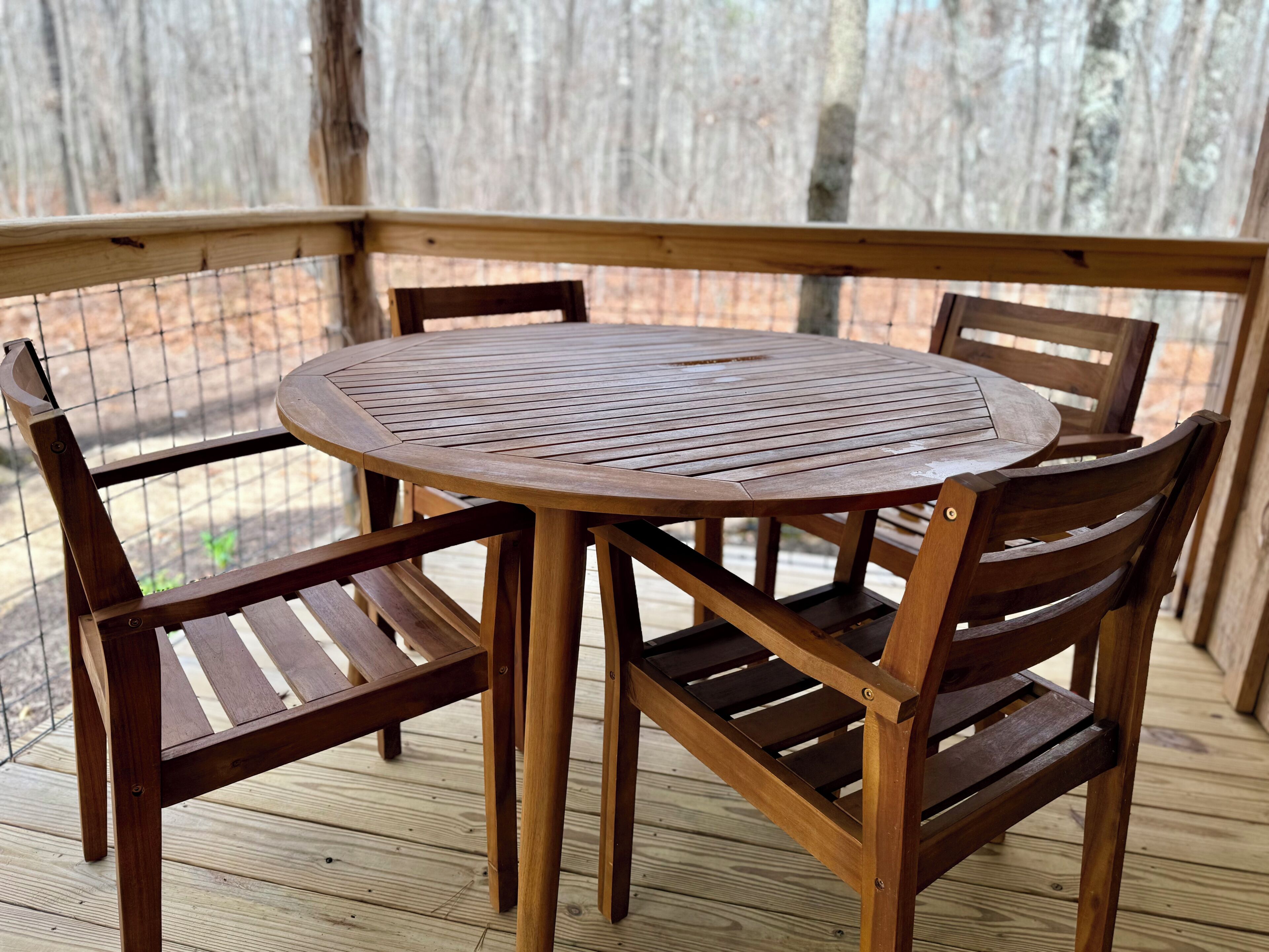 Una mesa redonda de madera con asientos de rejilla metálica, rodeada de sillas de madera sobre una terraza de madera.
