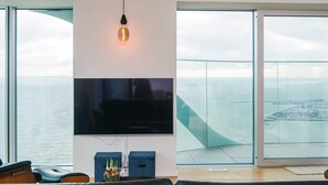 A modern living room with a large flat-screen TV mounted on a white wall, leather seating, and a view of the ocean through floor-to-ceiling glass doors. - 5 Star Holiday Home in Aarhus C (Aarhus)