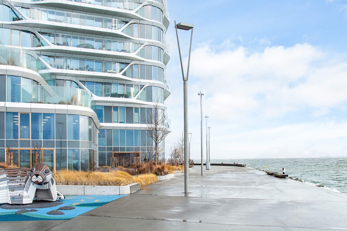Een modern gebouw met een gebogen gevel, uitzicht op het water en een betonpad met een blauw patroon.