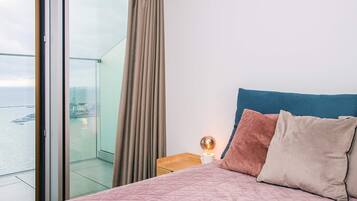 A hotel room with a large bed, a nightstand with a lamp, a view of the sea through a glass door, and a balcony.