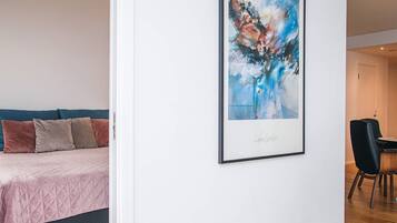 A bedroom with a bed, pillows, a framed abstract painting, and a dining area with chairs.