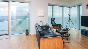 A modern living room with a large black leather sofa, a wooden coffee table, and a floor lamp. There is a tripod with a camera and a television on the wall. The room has large windows with a view of the ocean. - 5 Star Holiday Home in Aarhus C (Aarhus)