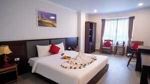 A hotel room with a neatly made bed, a bedside table with a lamp, a desk with a chair, and a window with curtains. - Flamant Boutique Hotel (Sa Pa)