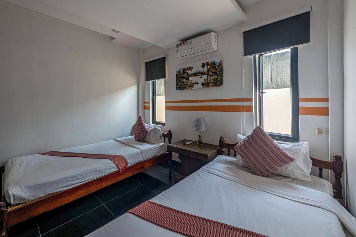 A hotel room with two single beds, a large bed, a window, an air conditioning unit, and a painting on the wall.