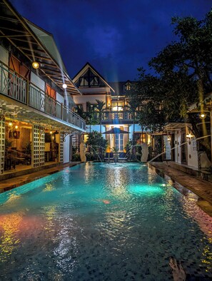 A pool area with a two-story building in the background, surrounded by trees and a well-lit balcony.