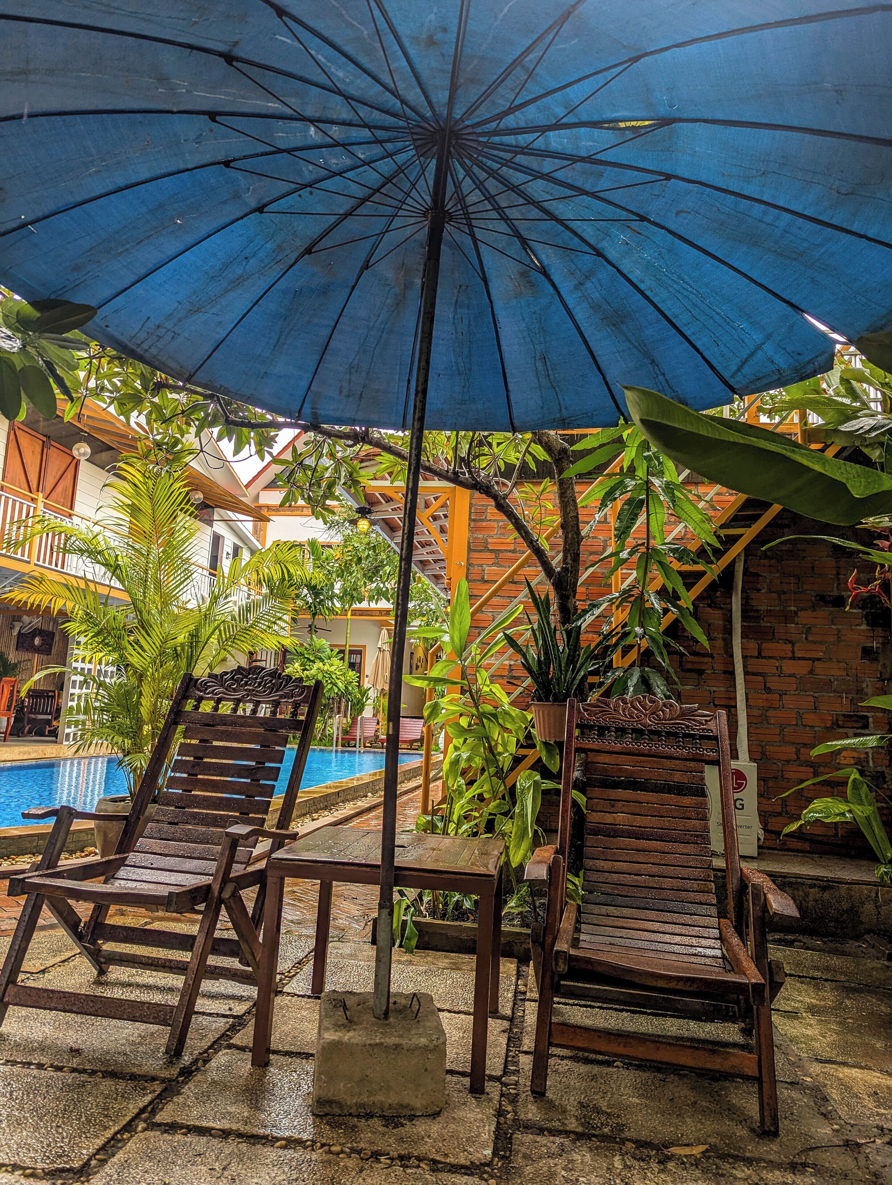 Un espace aménagé au bord de la piscine, avec deux chaises longues en bois sous un parasol bleu, entouré d’une végétation luxuriante et d  ’un mur en briques.