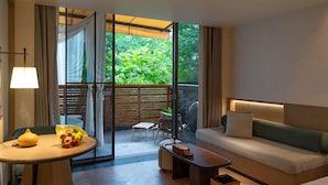 A modern hotel room with a sofa, a small table with a lamp, a chair, and a view of greenery through large windows. - Yueyin Chongqing Courtyard (Chongqing)