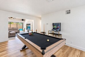 Ein Billardtisch mit Queue, Billardkugeln und einer Spielkonsole in einem modernen Raum, im Hintergrund ist eine Couch und ein weiterer Billardtisch zu sehen.