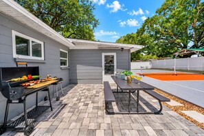 A patio area with a barbecue grill, a table with drinks, and a tennis court in the background.