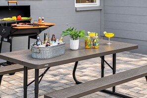 Un patio exterior con una parrilla, una mesa metálica con botellas, una planta en maceta y una bebida.