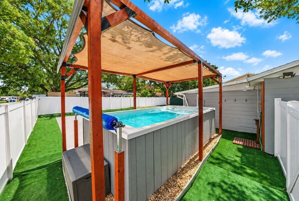 Un patio trasero con un jacuzzi, una pérgola de madera y césped artificial.