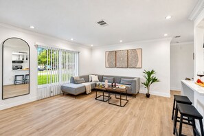 Una sala moderna con un sofá gris, una mesa de centro y un comedor con encimera blanca y taburetes de bar.