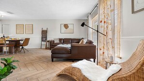 Ein modernes Wohnzimmer mit einem braunen Ledersofa, einem Esstisch mit Stühlen, einem Korbstuhl und einer weißen, flauschigen Decke.