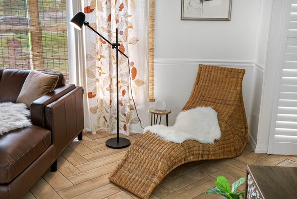 A cozy room with a brown leather sofa, a wicker chair with a white fur throw, a floor lamp, and a window with patterned curtains.