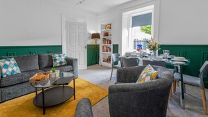 Een moderne woonkamer met een bank, fauteuil en salontafel, een eethoek met een tafel gedekt voor vier personen, en een boekenplank vol boeken.