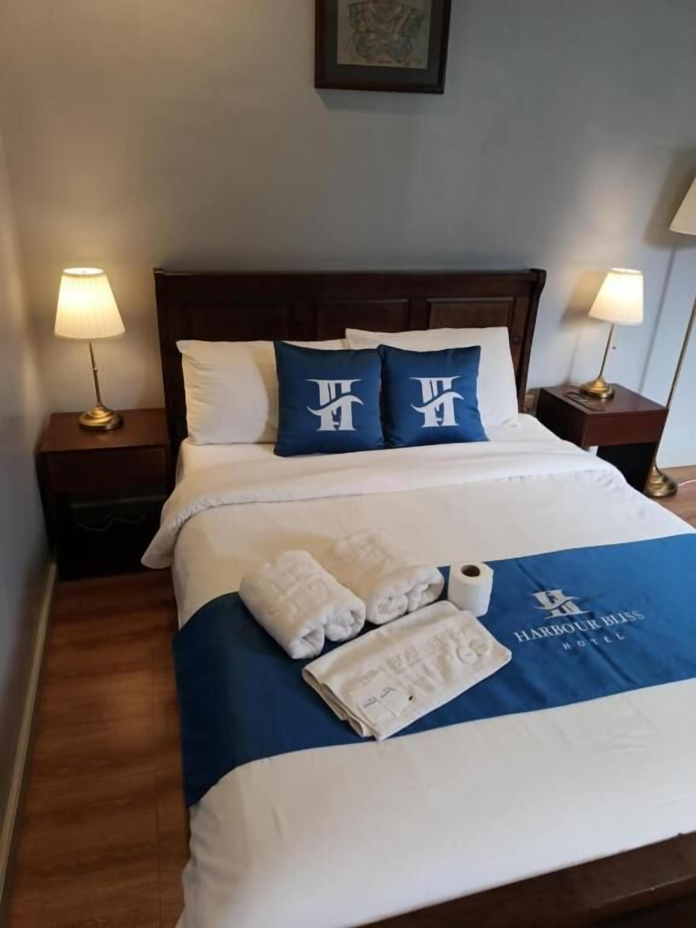 A hotel room with a neatly made bed, two pillows with a logo, a blue and white bedspread, two rolled towels, and a framed picture on the wall.