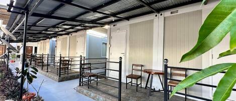 A covered outdoor seating area with tables and chairs, a stone-tiled floor, and a metal railing.