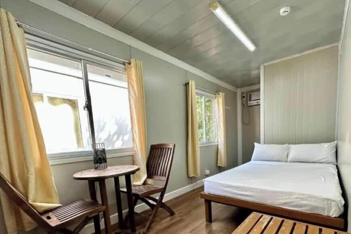 A room with a bed, a small table, a chair, and two windows with curtains.