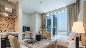 A modern hotel room with a dining area, kitchenette, and a large window offering a city view.