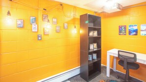 Ein Zimmer mit gelb gestrichenen Ziegelwänden, einem weißen Schreibtisch, einem schwarzen Bücherregal und gerahmten Bildern an der Wand.