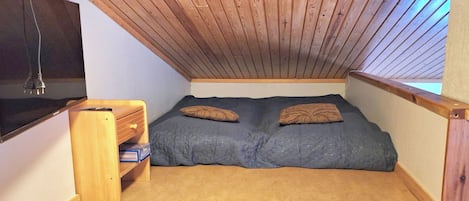 A small, sloped ceiling room with a bed, a shelf with books, and a TV mounted on the wall.