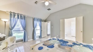 Un dormitorio amplio con una cama grande, un escritorio y una silla. Hay dos ventanas con cortinas azules, un ventilador de techo y una puerta que conduce a otra habitación.