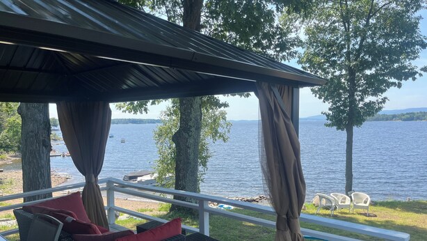 A covered outdoor seating area with wicker furniture and red cushions, overlooking a lake with boats and trees.