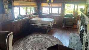 A rustic cabin interior with a wooden dining table, a built-in bench, a fireplace, and a large window providing natural light.