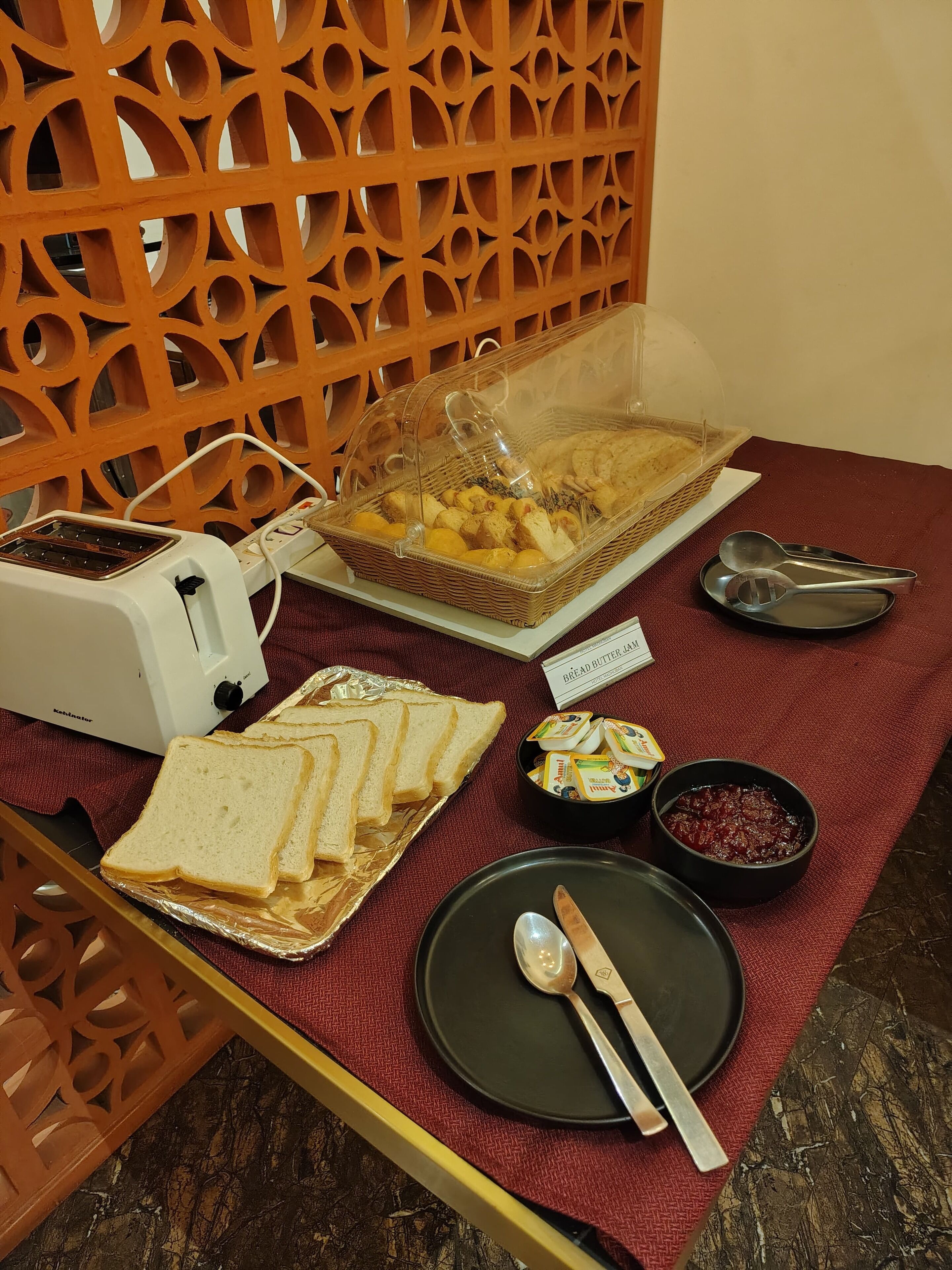 Une table dressée pour le petit-déjeuner, avec un grille-pain, du pain, un panier de viennoiseries et des condiments, le tout posé sur une nappe rouge.