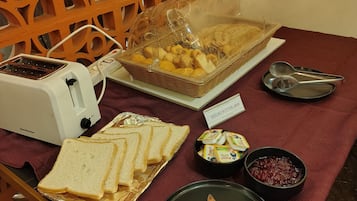 Une table dressée pour le petit-déjeuner, avec un grille-pain, du pain, un panier de viennoiseries et des condiments, le tout posé sur une nappe rouge.