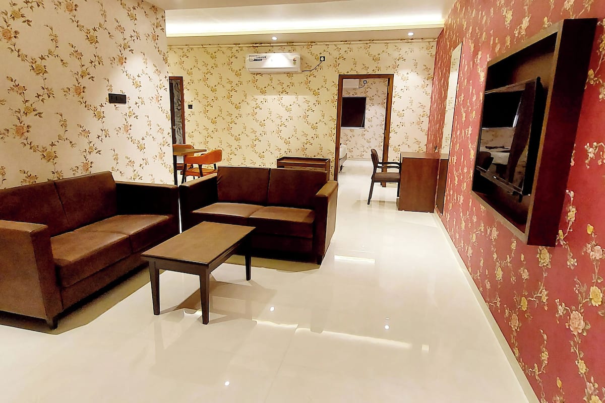Una sala de estar con sofá, sillón y una mesa de centro. Hay un televisor montado en la pared y una puerta que conduce a otra habitación.