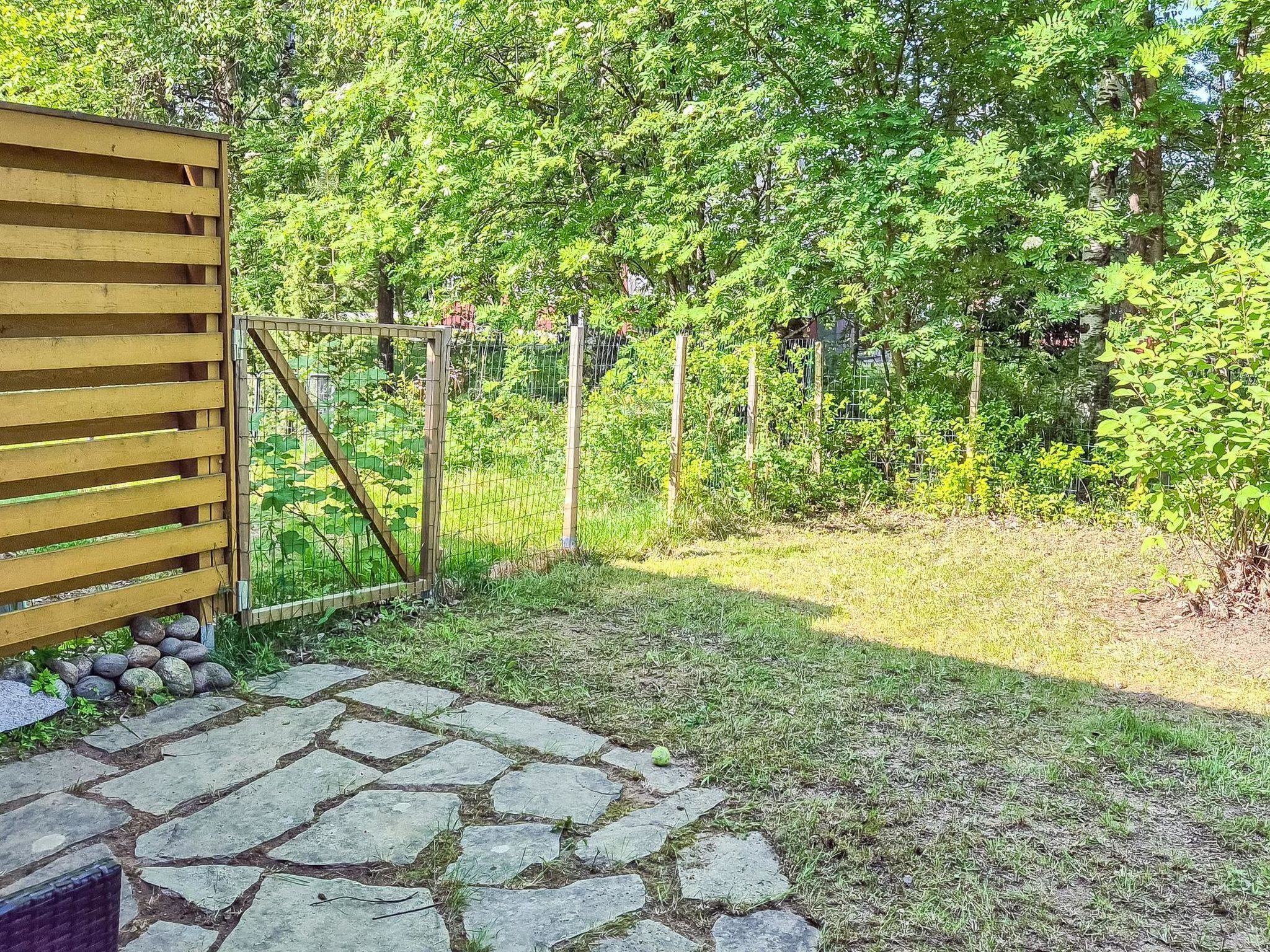 Ein Steinweg führt zu einem hölzernen Tor, umgeben von üppiger Vegetation und einem eingezäunten Bereich.