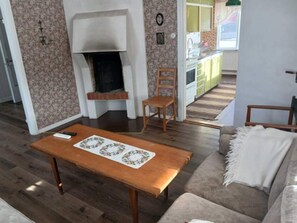 A living room with a fireplace, a sofa, a wooden table, and a kitchen in the background.