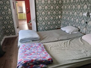 A room with two beds, a patterned wallpaper, and a visible kitchen area in the background.