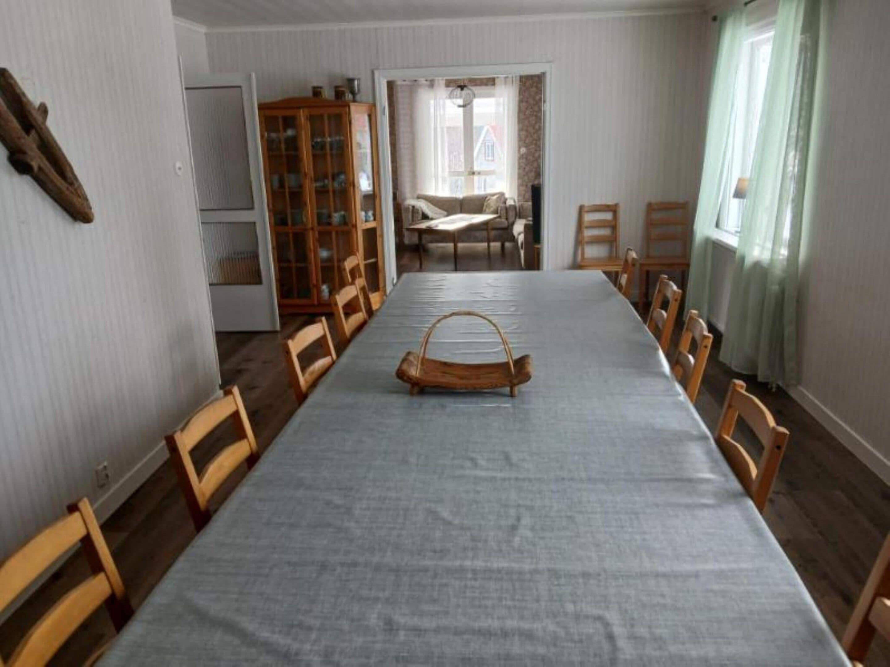 Comedor con una larga mesa, sillas de madera, un aparador con puertas de vidrio y vista a un dormitorio a través de una puerta abierta.