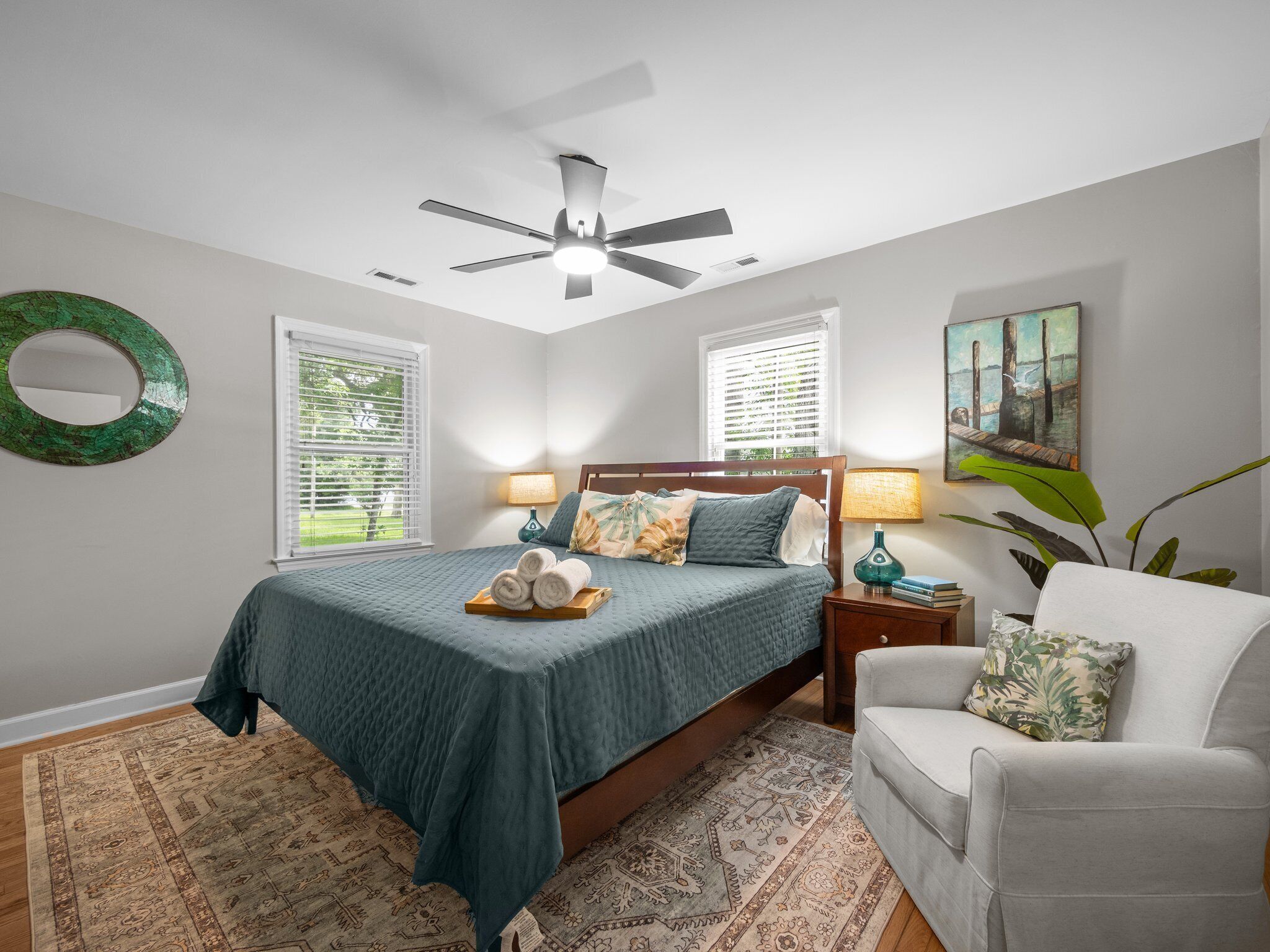A bedroom with a bed, a ceiling fan, two bedside lamps, a chair, a rug, and a painting on the wall.