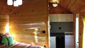 Interior de una cabaña de madera con una cama, un rincón de cocina y un ventilador de techo.