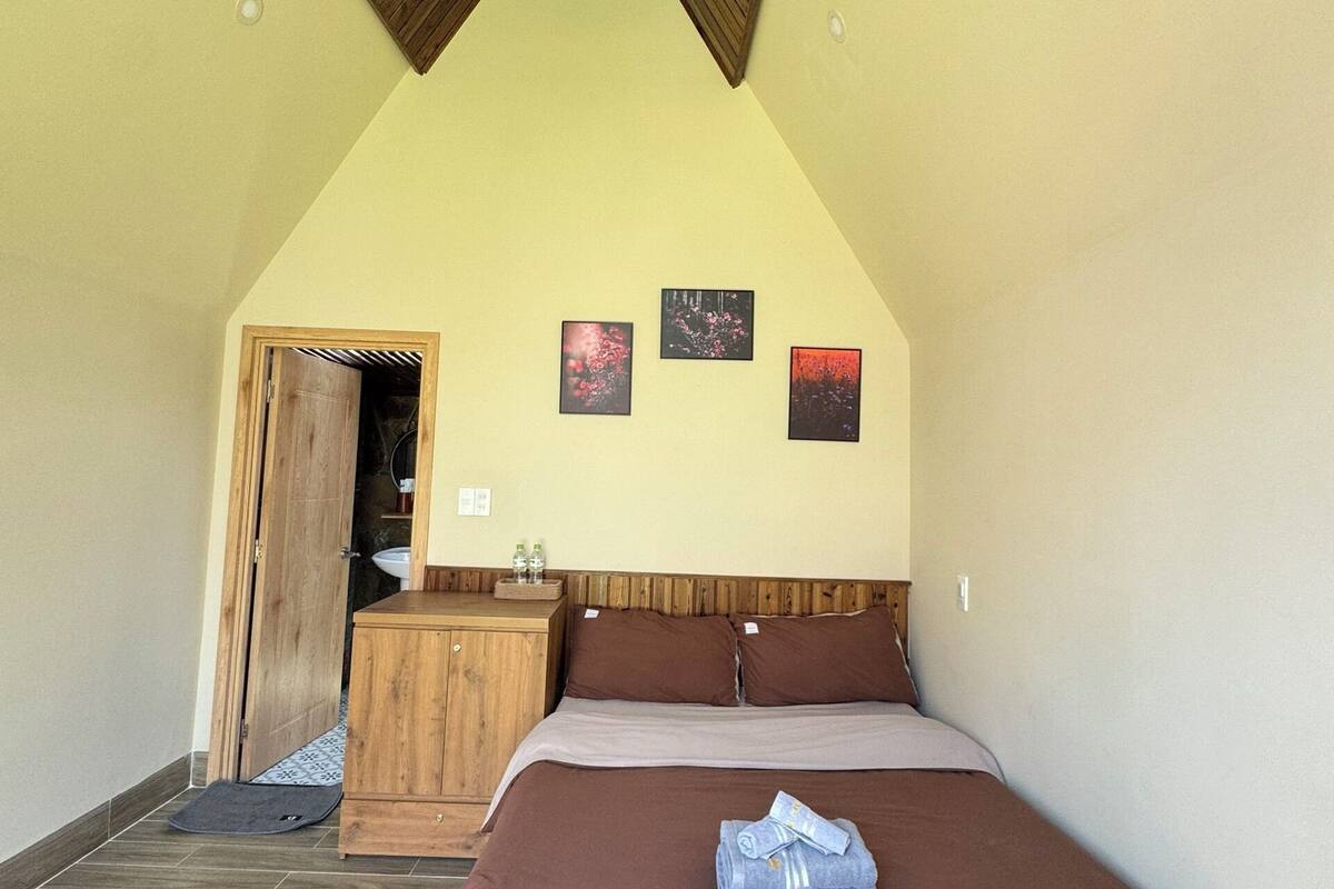 A bedroom with a sloped ceiling, a wooden headboard, a bed with brown bedding, a small wooden dresser, and three framed pictures on the wall.