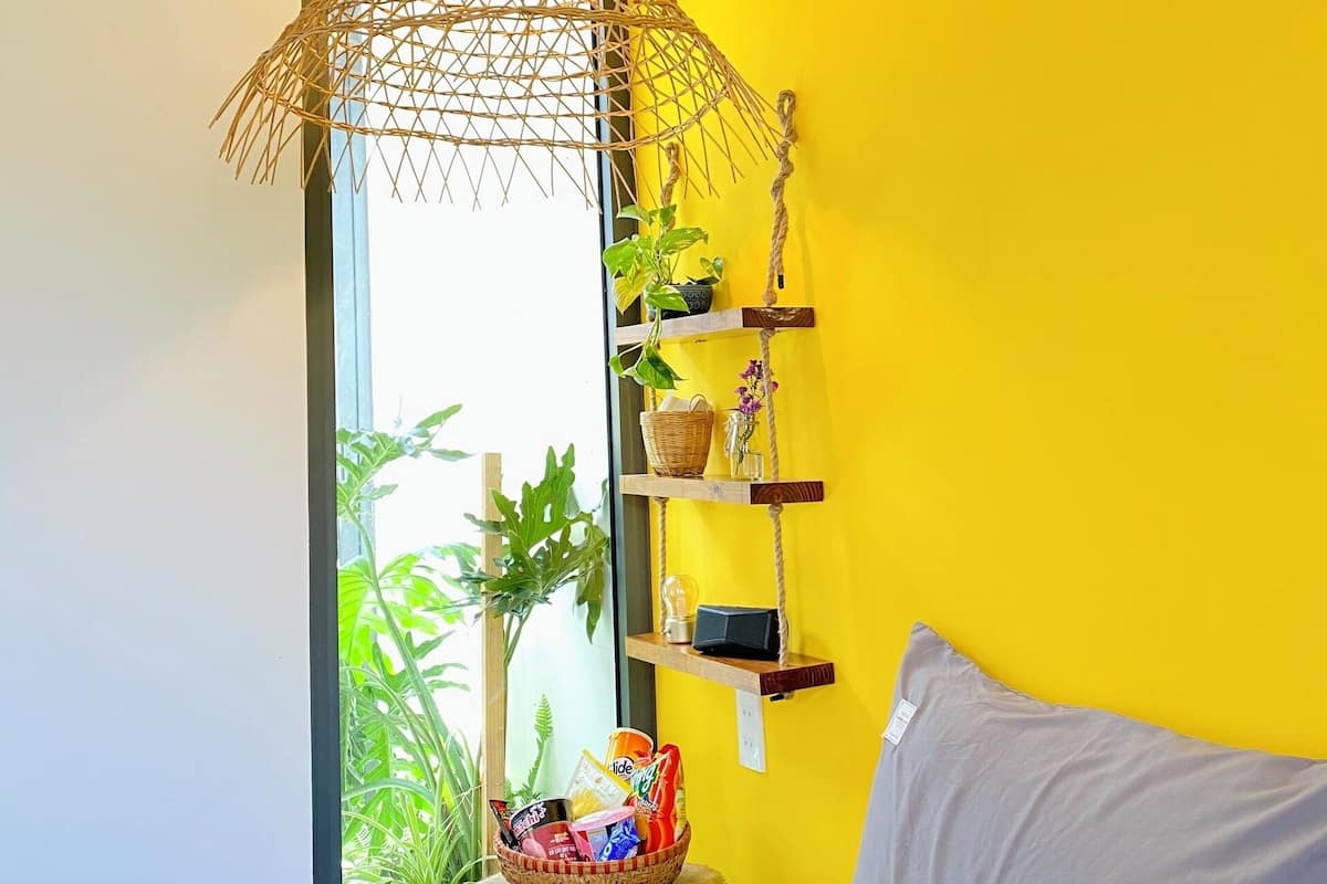 A room with a bed, a hanging light fixture, a shelf with plants and items, and a window with greenery outside.