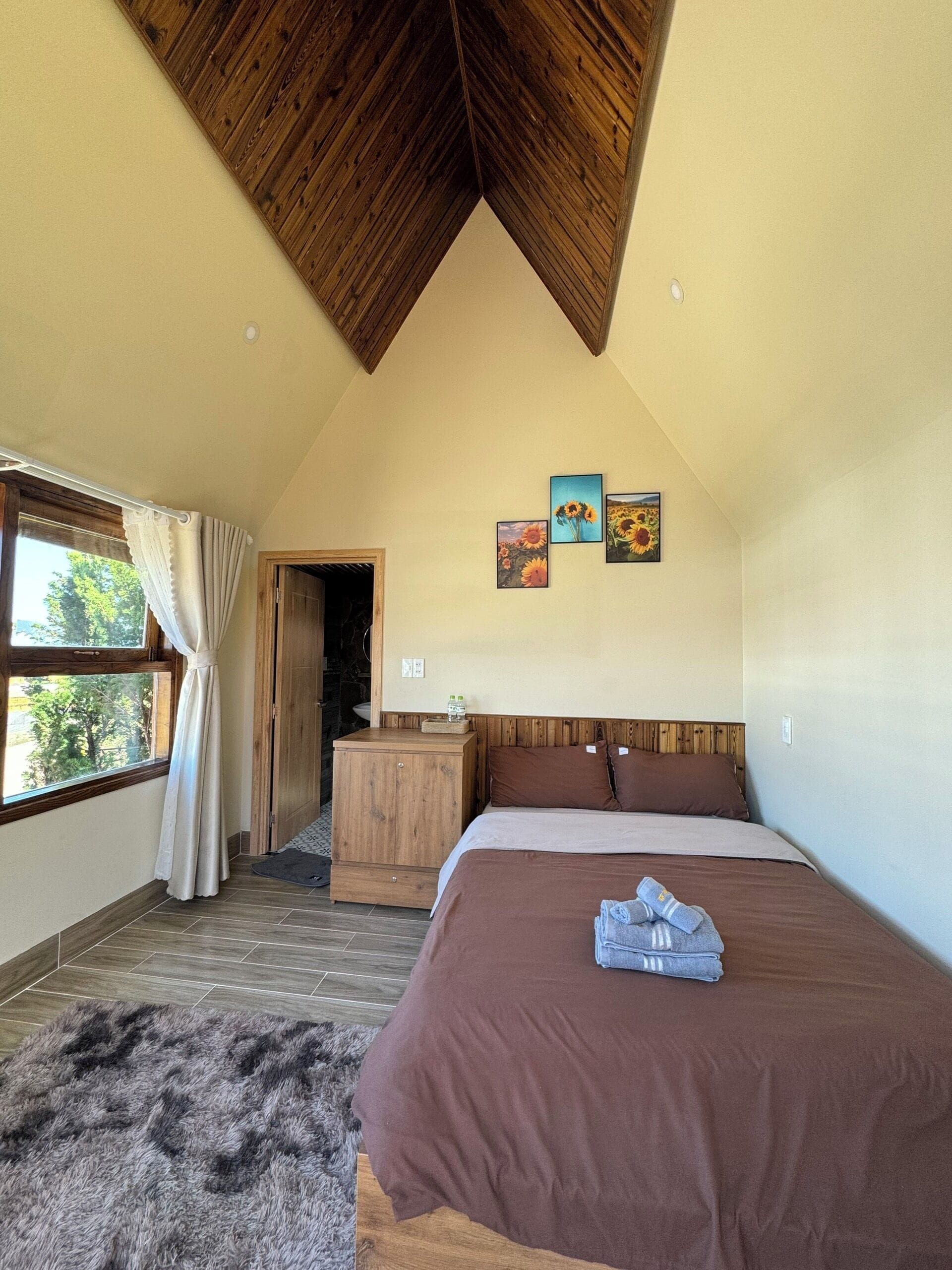 Une chambre avec un plafond en pente, un lit avec une literie marron, une table de chevet en bois, une petite armoire et une fenêtre avec des rideaux.