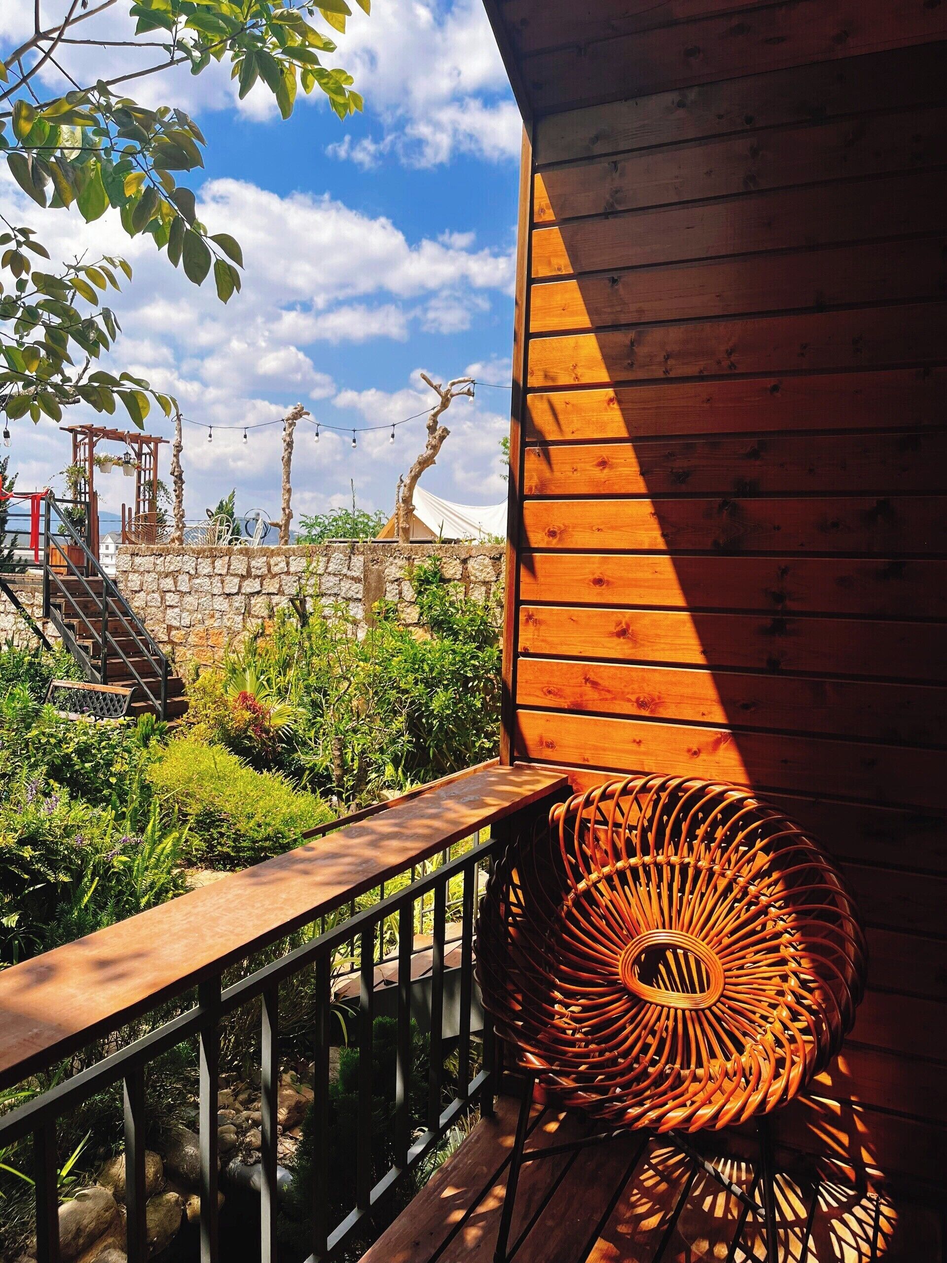 Une terrasse en bois avec un fauteuil en osier, entourée d’une végétation luxuriante et d’un mur de pierre.