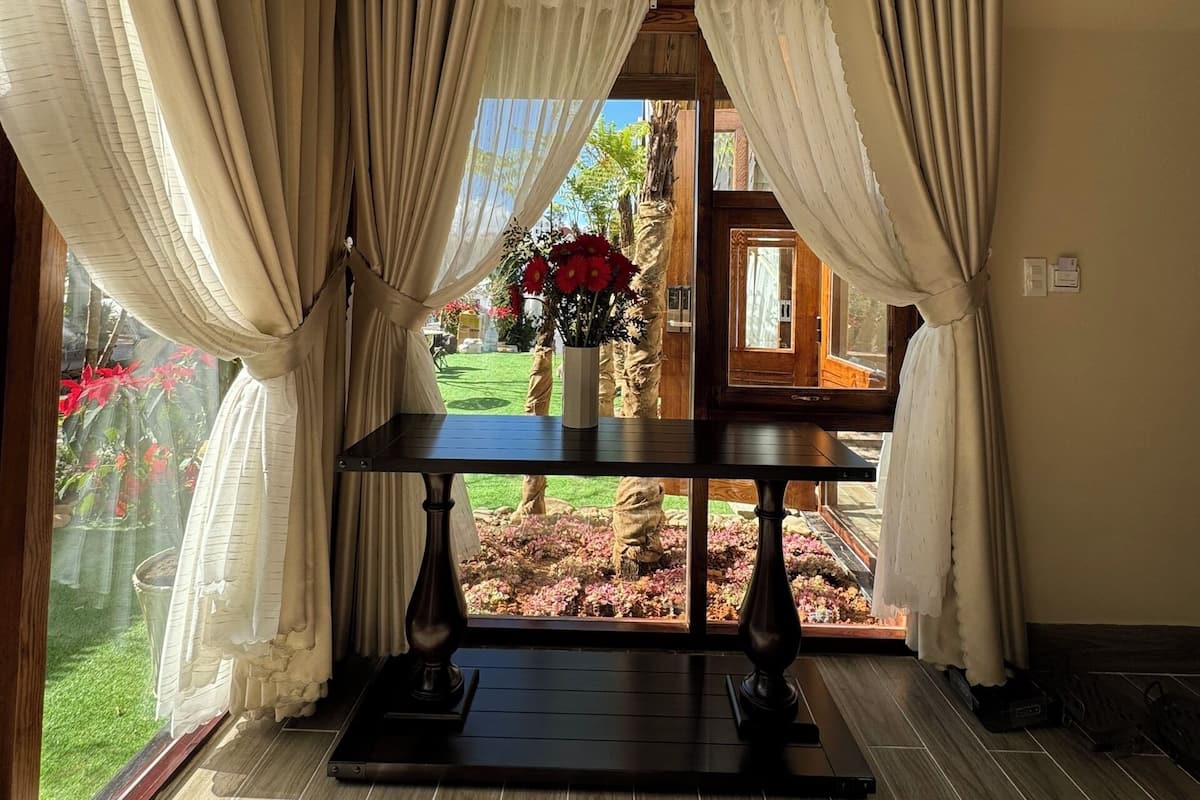 A room with a view of a garden through large windows, a table with a vase of flowers, and a wooden floor.