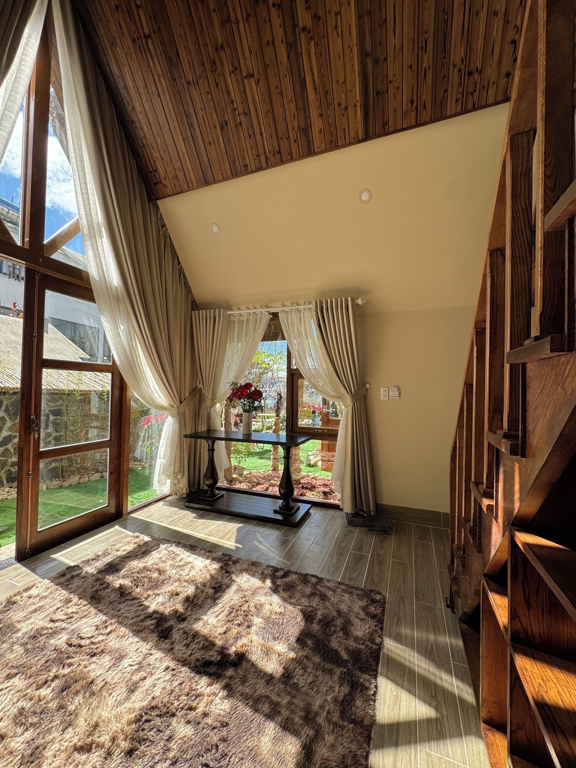 Une pièce spacieuse avec un plafond en bois, de grandes fenêtres dotées de rideaux, un escalier en bois et une vue sur un jardin.