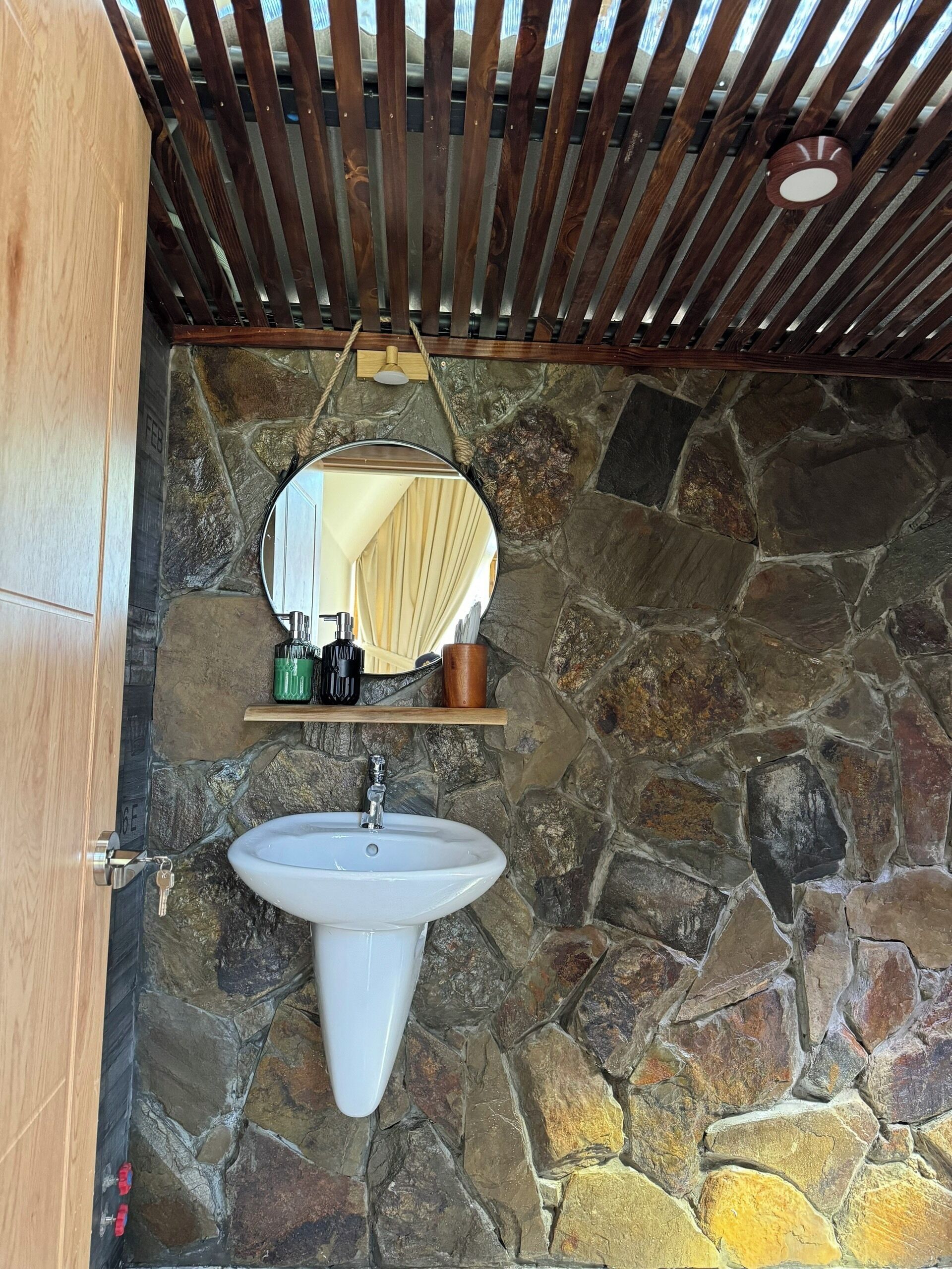 Un baño con pared de piedra, puerta de madera, lavamanos blanco, espejo y repisa con artículos de aseo.