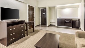 Una sala moderna con televisor de pantalla plana, muebles de madera oscura, un sofá color beige y una cocina equipada con microondas y refrigerador.