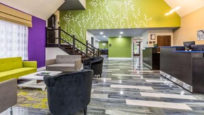 A hotel lobby with a reception desk, seating area, staircase, and a green wall with white tree designs. - Travelodge by Wyndham Flowood (Flowood)