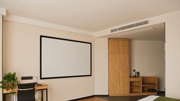 A modern hotel room with a wooden desk, a chair, a bed with a green blanket, a wardrobe, and a large blank wall. - Greentree Inn Nanjing Baijiahu Express Hotel (Nanjing)