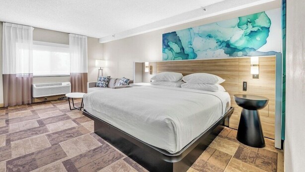 Una habitación de hotel moderna con una cama grande, una obra de arte colgada en la pared, una mesita de noche y un ventanal con cortinas.