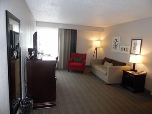 A hotel room with a desk, a red chair, a sofa, a lamp, and framed pictures on the wall. - Wingate by Wyndham Jacksonville FL (Jacksonville)